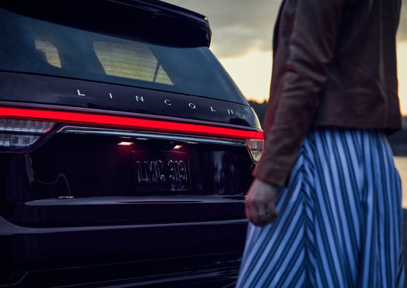 A person is shown near the rear of a 2024 Lincoln Aviator® SUV as the Lincoln Embrace illuminates the rear lights | West Point Lincoln in Houston TX